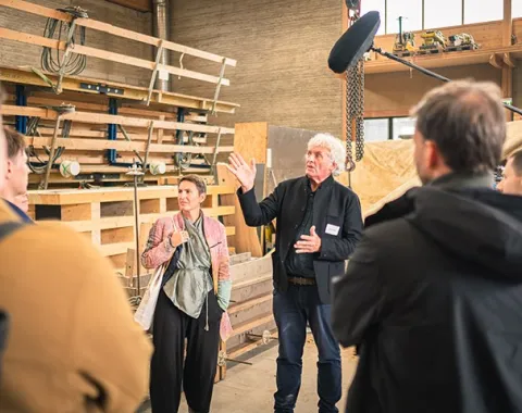 Eine Gruppe von Menschen steht in einer Werkstatt. Ein Mann spricht gestikulierend zu den Anwesenden. Im Hintergrund sind Holzregale und Werkzeuge zu sehen.