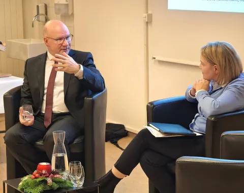 Zwei Personen sitzen in Sesseln und führen ein Gespräch. Der Mann links trägt einen Anzug und hält ein Glas, während er gestikuliert. Die Frau rechts hält ein Notizbuch. Im Vordergrund steht ein Tisch mit einer Karaffe und einem weihnachtlichen Gesteck.