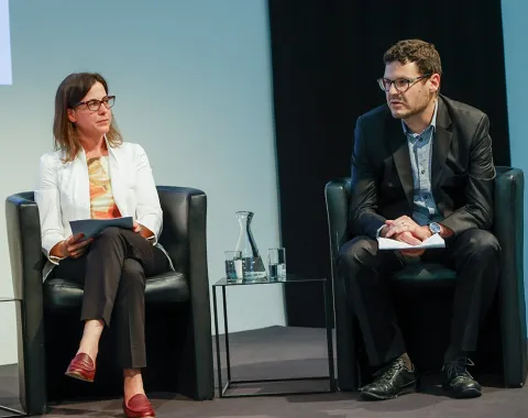 Zwei Personen sitzen auf einer Bühne in schwarzen Sesseln. Die Frau links trägt eine weiße Jacke und hält Papiere, der Mann rechts trägt einen dunklen Anzug. Zwischen ihnen steht ein Wasserglas auf einem kleinen Tisch.
