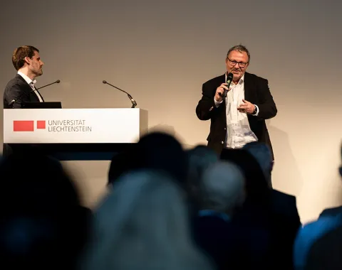 Zwei Männer stehen auf einer Bühne bei einer Veranstaltung an der Universität Liechtenstein. Einer spricht in ein Mikrofon, während der andere neben einem Rednerpult mit dem Logo der Universität steht. Im Vordergrund sind unscharf die Köpfe des Publikums zu sehen.