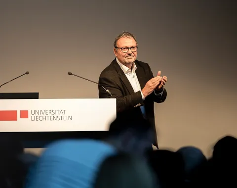 Ein Mann steht an einem Rednerpult der Universität Liechtenstein und applaudiert. Im Hintergrund sind Mikrofone sichtbar.