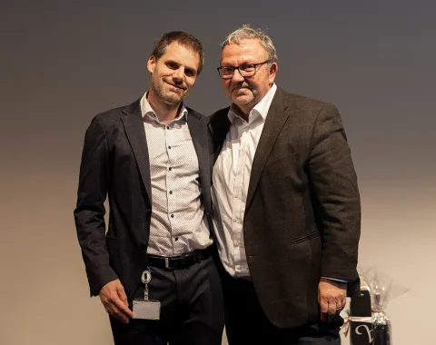 Zwei Männer stehen nebeneinander und lächeln in die Kamera. Sie tragen Anzüge und befinden sich in einem Innenraum mit neutralem Hintergrund.