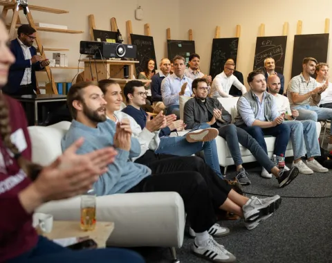 Eine Gruppe von Menschen sitzt in einem Raum und applaudiert. Im Hintergrund sind Tafeln und technische Geräte zu sehen. Die Atmosphäre wirkt konzentriert und feierlich.