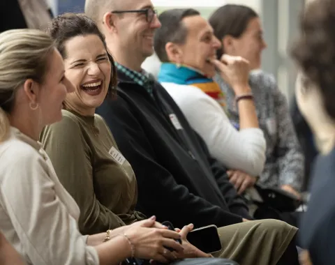 Eine Gruppe von Menschen sitzt lachend in einem Veranstaltungsraum. Sie wirken entspannt und fröhlich, während sie einem Vortrag oder einer Präsentation folgen.