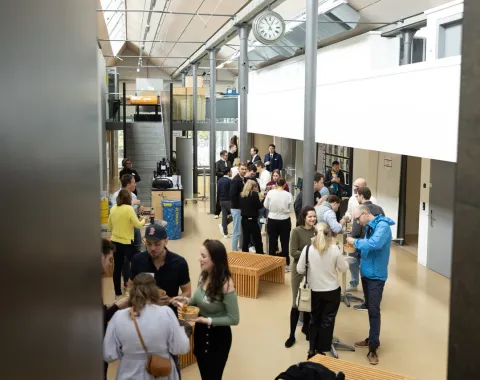 Eine Gruppe von Menschen versammelt sich in einem modernen, lichtdurchfluteten Innenraum der Universität Liechtenstein. Sie stehen in kleinen Gruppen zusammen und unterhalten sich. Im Hintergrund sind Treppen und eine Uhr zu sehen.