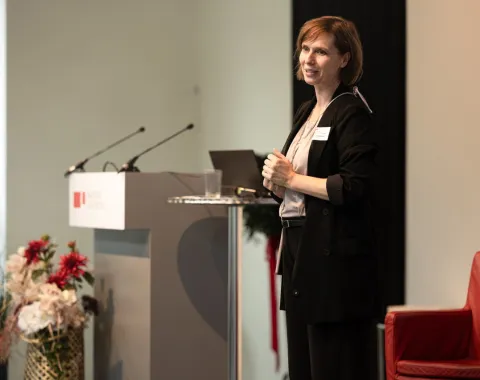 Eine Person steht auf einer Bühne neben einem Rednerpult mit Mikrofonen. Im Hintergrund sind Blumenarrangements und ein roter Stuhl zu sehen.