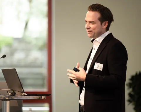 Ein Redner im Anzug hält einen Vortrag auf einer Veranstaltung an der Universität Liechtenstein. Im Hintergrund sind ein Fenster und Pflanzen zu sehen.