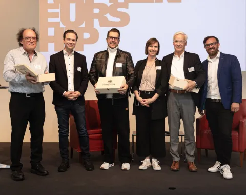 Sechs Personen stehen auf einer Bühne und halten Geschenke in den Händen. Im Hintergrund ist ein Bildschirm mit dem Wort "Entrepreneurship" zu sehen. Die Personen lächeln und tragen Namensschilder.