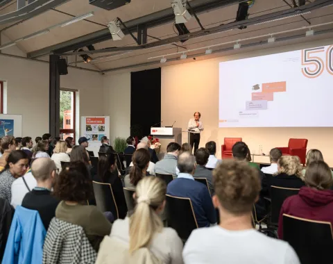 Ein Vortragender steht auf einer Bühne vor einem Publikum in einem modernen Veranstaltungsraum. Auf der Leinwand hinter ihm ist eine Präsentation mit der Zahl "50" zu sehen. Die Zuhörer sitzen aufmerksam in Reihen.