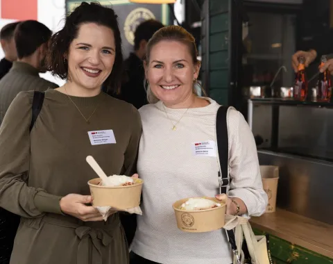 Zwei lächelnde Personen halten Schalen mit Essen in den Händen, während sie bei einer Feier an der Universität Liechtenstein stehen. Im Hintergrund ist ein Essensstand zu sehen.