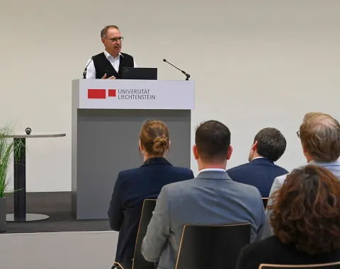 Ein Redner steht an einem Podium mit dem Logo der Universität Liechtenstein und spricht zu einem sitzenden Publikum. Im Hintergrund sind Pflanzen zu sehen.