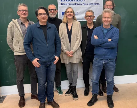 Eine Gruppe von acht Personen steht vor einem grünen Hintergrund. Sie lächeln in die Kamera. Im Hintergrund hängt ein Banner mit der Aufschrift "Erasmus+ Neue Perspektiven, Neue Horizonte".