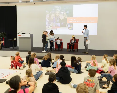 Ein Vortragssaal mit Kindern, die auf dem Boden sitzen und einem Vortragenden zuhören. Einige Kinder stehen auf der Bühne und nehmen an einer interaktiven Aktivität teil. Im Hintergrund ist eine Präsentation zu sehen.