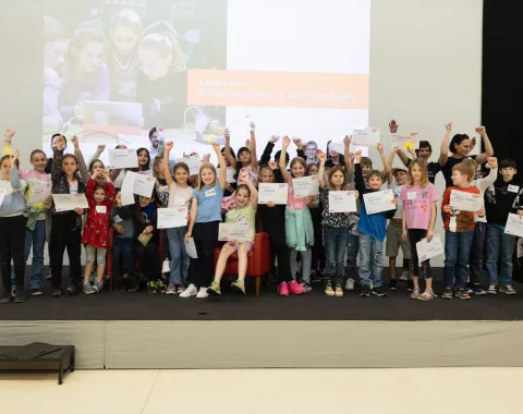 Gruppe von Kindern auf einer Bühne, die stolz Zertifikate hochhalten, im Rahmen der Kinder-Uni an der Universität Liechtenstein. Im Hintergrund ein Bildschirm mit der Aufschrift „Entdeckungsreise ins Reich der Regeln“.