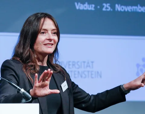 Eine Frau hält einen Vortrag bei einer Konferenz an der Universität Liechtenstein. Sie trägt ein Mikrofon und gestikuliert mit den Händen. Im Hintergrund ist ein Banner mit dem Datum der Veranstaltung und dem Logo der Universität zu sehen.
