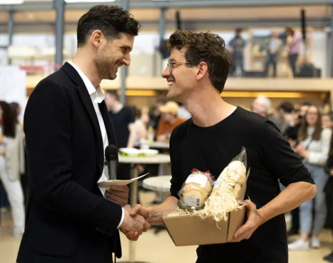 Zwei Männer schütteln sich die Hand bei einer Veranstaltung in einem Innenraum. Einer hält ein Mikrofon, der andere ein Geschenkpaket. Im Hintergrund sind weitere Personen unscharf zu erkennen.