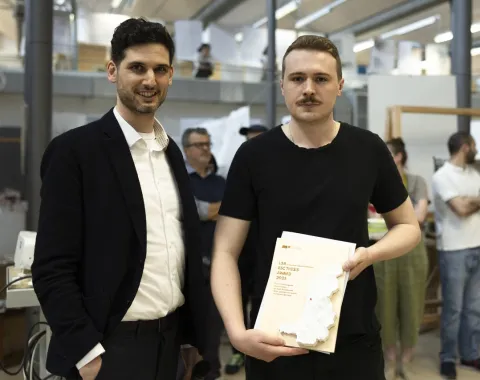 Zwei Personen stehen in einem Innenraum. Eine Person hält ein Zertifikat oder eine Auszeichnung in der Hand. Im Hintergrund sind weitere Personen und architektonische Modelle zu sehen. Die Szene wirkt wie eine Preisverleihung oder ein Event an einer Universität.