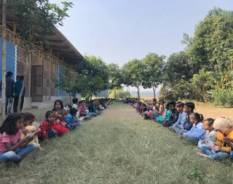 Kinder sitzen in zwei Reihen auf einer Wiese vor einem Schulgebäude in Indien. Sie sind von Bäumen umgeben und scheinen an einer Aktivität teilzunehmen. Die Szene vermittelt Gemeinschaft und Bildung im Freien.