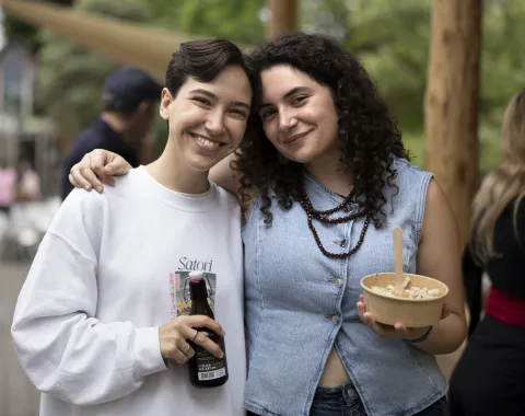 Zwei lächelnde Personen stehen eng beieinander auf einem Sommerfest. Eine Person hält eine Getränkeflasche, die andere eine Schüssel mit Essen. Im Hintergrund sind unscharfe Bäume und weitere Gäste zu sehen.