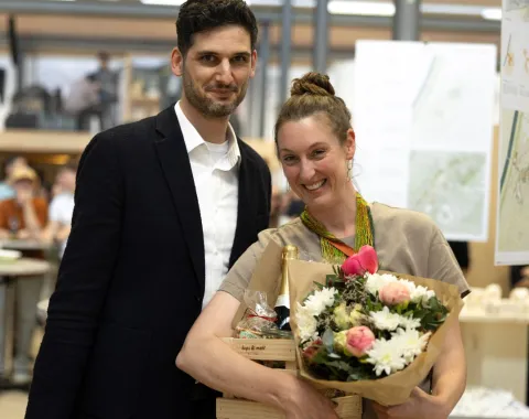 Zwei Personen stehen lächelnd in einem Innenraum. Eine Person hält einen Blumenstrauß und eine Holzkiste mit Geschenken. Im Hintergrund sind unscharf architektonische Pläne und weitere Personen zu erkennen.