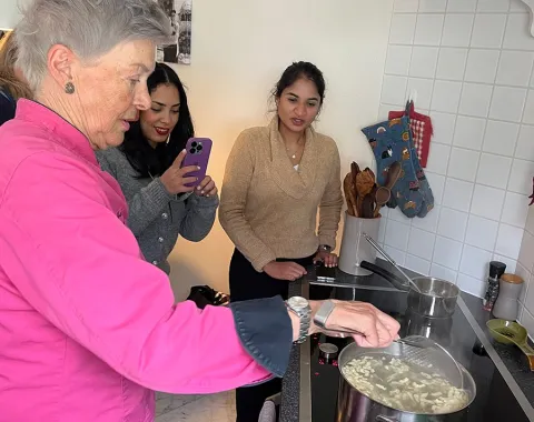 Eine Gruppe von Menschen steht in einer Küche. Eine Person in einem pinken Oberteil kocht an einem Herd, während zwei andere Personen zuschauen und eine von ihnen mit einem Smartphone fotografiert. Auf dem Herd steht ein Topf mit kochendem Wasser.
