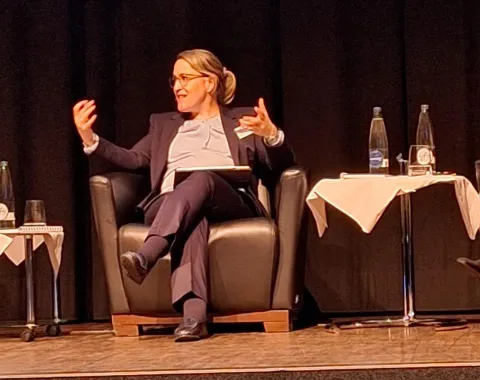 Eine Person sitzt auf einem Podium in einem Sessel und gestikuliert während einer Diskussion. Neben ihr stehen Tische mit Wasserflaschen und Gläsern. Die Szene findet in einem Konferenzsaal statt.