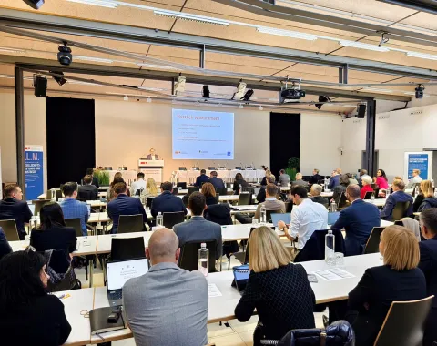 Ein Auditorium voller Menschen sitzt an Tischen und hört einem Vortrag zu. Auf einer Leinwand vorne sind Präsentationsfolien zu sehen. Die Veranstaltung findet in einem modernen, gut beleuchteten Raum statt.