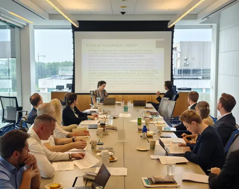 Ein Konferenzraum mit mehreren Personen, die an einem langen Tisch sitzen und einer Präsentation folgen. Im Hintergrund ist eine große Leinwand mit projiziertem Text zu sehen. Die Teilnehmer sind in eine Diskussion vertieft.