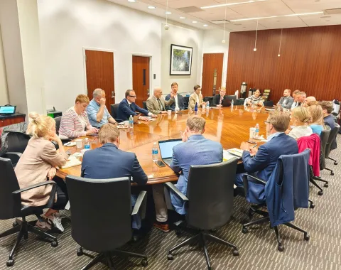 Eine Gruppe von Studierenden und Dozenten sitzt in einem Konferenzraum an einem großen, ovalen Tisch. Sie sind in einer Besprechung vertieft, umgeben von Büromöbeln und moderner Einrichtung.