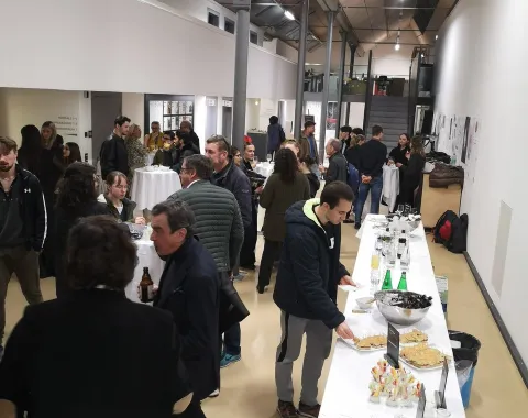 Eine Gruppe von Menschen steht in einem modernen Veranstaltungsraum, unterhält sich und genießt ein Buffet mit Getränken und Snacks. Im Hintergrund sind hohe Decken und eine Uhr sichtbar.