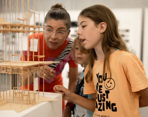 Eine Frau und zwei Kinder betrachten ein detailliertes Architekturmodell aus Holz. Die Szene findet in einem Ausstellungsraum statt, der Teil des Tags der offenen Tür an der Universität Liechtenstein ist.