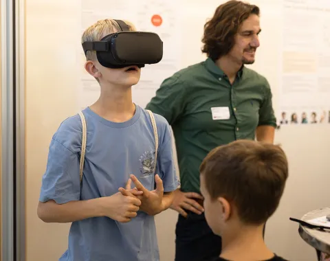 Ein Junge trägt eine VR-Brille und erkundet virtuelle Inhalte. Im Hintergrund steht ein Mann, der lächelnd zusieht. Weitere Personen sind teilweise sichtbar. Die Szene spielt in einem Raum mit Informationspostern an der Wand.