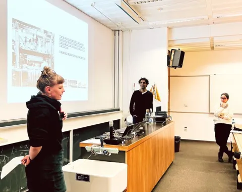 Eine Präsentation in einem Universitätsseminarraum: Drei Personen stehen vor einer Leinwand, auf der eine Folie projiziert wird. Eine Person spricht, während die anderen zuhören. Der Raum ist mit Tischen und einer Tafel ausgestattet.