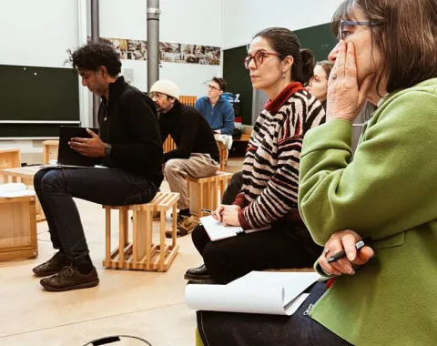 Eine Gruppe von Personen sitzt aufmerksam in einem Seminarraum. Sie sind auf Holzhockern platziert und scheinen einer Präsentation oder Diskussion zu folgen. Im Hintergrund sind Tafeln und Poster zu sehen. Die Szene vermittelt eine konzentrierte Lernatmosphäre.