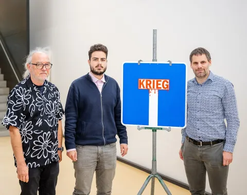 Drei Männer stehen neben einem blauen Verkehrsschild mit der Aufschrift "Krieg", das symbolisch eine Sackgasse darstellt. Das Bild wurde bei der Veranstaltung "Verkehrsschilder der Gerechtigkeit" an der Universität Liechtenstein aufgenommen.