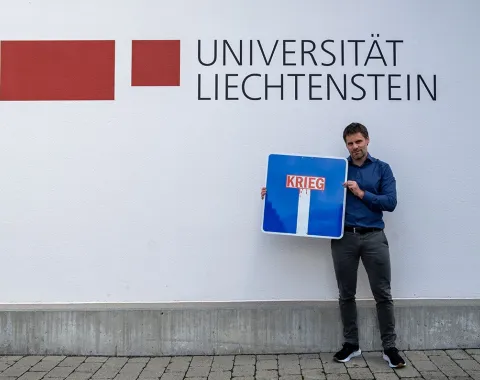 Ein Mann steht vor einer Wand mit dem Logo der Universität Liechtenstein und hält ein Verkehrsschild mit der Aufschrift „Krieg“ in einer Sackgasse.