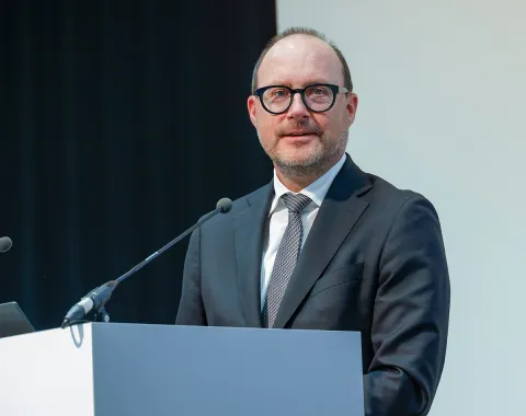 Ein Mann in Anzug und Krawatte steht an einem Rednerpult und spricht in ein Mikrofon. Der Hintergrund ist schlicht und professionell gestaltet.