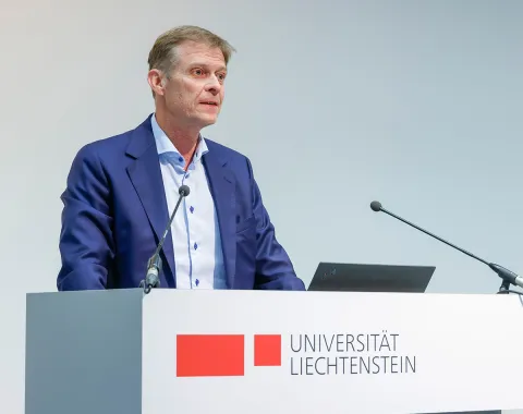 Ein Mann steht an einem Rednerpult mit dem Logo der Universität Liechtenstein und spricht in ein Mikrofon.