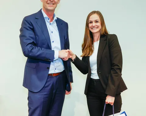 Zwei Personen stehen auf einer Bühne und schütteln sich die Hand. Die Frau hält eine Geschenktasche mit dem Logo der Universität Liechtenstein. Beide lächeln und tragen formelle Kleidung.