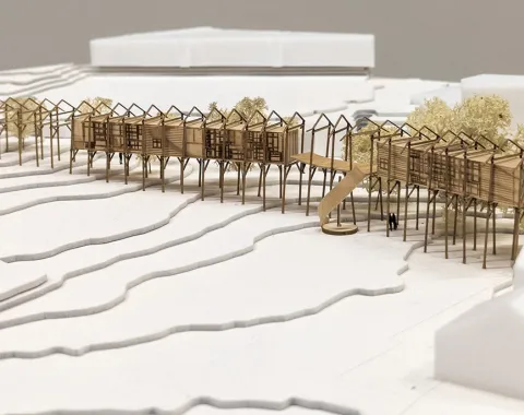 Architekturmodell eines linearen Gebäudekomplexes auf Stelzen, eingebettet in eine Landschaft mit Höhenlinien. Das Modell zeigt eine innovative Wiederverwendung von Bauteilen eines alten Strickbaus, umgeben von stilisierten Bäumen und Gebäuden.