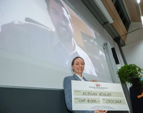 Eine Person hält einen großen Scheck mit der Aufschrift "Adrian Vogler, CHF 4,000, 29.01.2024" in einem Vortragsraum. Im Hintergrund ist eine Projektion eines lächelnden Mannes auf einer Leinwand zu sehen.