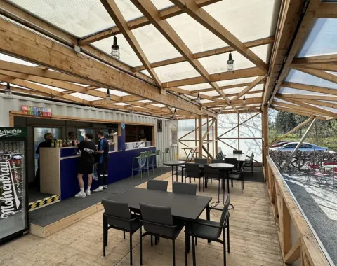 Ein moderner, überdachter Raum mit Holzkonstruktion und transparentem Dach. Links befindet sich eine Bar mit Personen, die bedient werden. Rechts stehen Tische und Stühle für Besucher bereit. Die Umgebung wirkt einladend und funktional für Veranstaltungen oder Treffen.