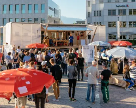 Eine Menschenmenge versammelt sich vor einer modularen Plattform im Freien, umgeben von modernen Gebäuden. Die Plattform dient als Veranstaltungsort mit Rednern und Sitzgelegenheiten. Bunte Sonnenschirme bieten Schatten. Die Szene zeigt eine lebendige, urbane Umgebung, die für Austausch und Beteiligung genutzt wird.