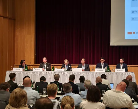 Ein Podium mit sechs Personen sitzt vor einem Publikum in einem Konferenzsaal. Auf dem Tisch stehen Namensschilder und Wasserflaschen. Im Hintergrund ist ein roter Vorhang und ein Teil einer Präsentationsleinwand zu sehen.