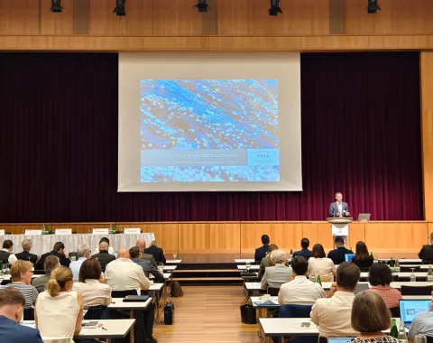 Ein Vortragssaal mit Teilnehmern, die an Tischen sitzen und einem Redner auf der Bühne zuhören. Eine große Leinwand zeigt eine Präsentation. Der Raum ist gut besucht und die Atmosphäre wirkt professionell.