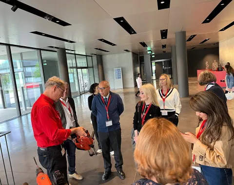 Eine Gruppe von Menschen steht in einem modernen Gebäude und hört einer Person zu, die etwas erklärt. Alle tragen Namensschilder und einige haben rote Lanyards. Im Hintergrund sind große Fenster und moderne Architektur zu sehen.
