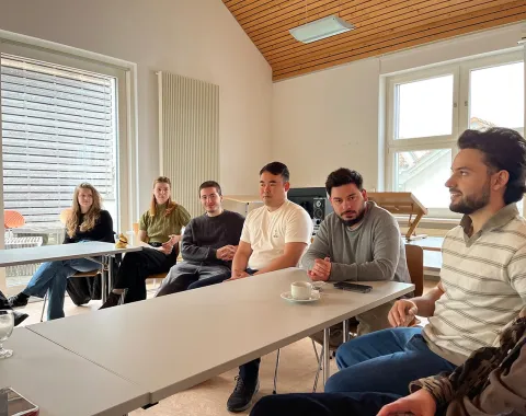 Eine Gruppe von Menschen sitzt in einem Besprechungsraum an Tischen. Sie sind in ein Gespräch vertieft. Im Hintergrund sind große Fenster und eine Holzdecke zu sehen.