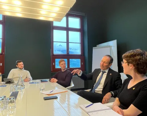 Ein Mann im Anzug spricht mit drei Personen an einem Konferenztisch in einem Raum mit großen Fenstern. Auf dem Tisch liegen Notizbücher und Gläser.