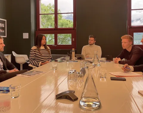 Eine Gruppe von Personen sitzt an einem Konferenztisch in einem Raum mit großen Fenstern. Sie diskutieren in einer formellen Umgebung. Auf dem Tisch stehen Wassergläser und Karaffen. Im Hintergrund sind grüne Bäume und ein bewölkter Himmel zu sehen.