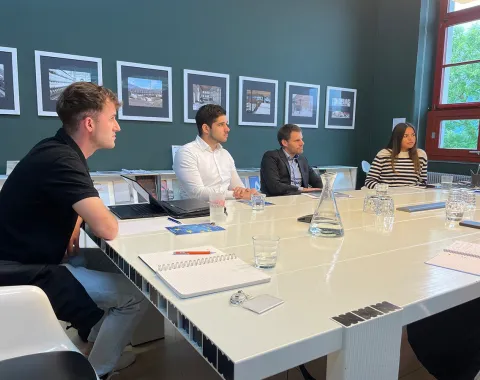 Eine Gruppe von vier Personen sitzt an einem weißen Konferenztisch in einem Raum mit grüner Wand und mehreren gerahmten Bildern. Auf dem Tisch liegen Notizbücher, Stifte und Gläser. Die Personen wirken konzentriert und diskutieren möglicherweise ein Thema. Im Hintergrund sind große Fenster zu sehen, die Tageslicht hereinlassen.